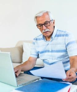 An old man sitting on a white sofa holding a document in his hands and working on a laptop
